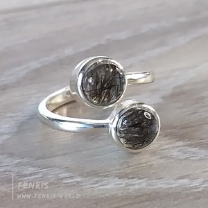 May include: A silver ring with two round, clear stones with black inclusions. The stones are set in silver bezels and the ring is open on one side.