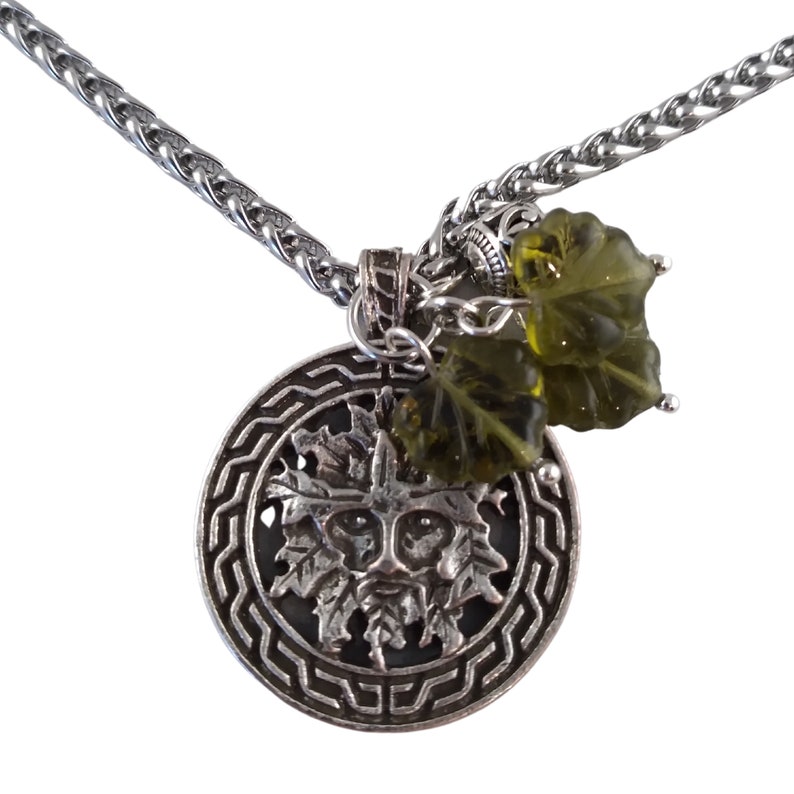 May include: A silver necklace with a round pendant featuring a green man face surrounded by a Celtic knot design. The pendant is attached to a silver chain with two green glass leaf charms.