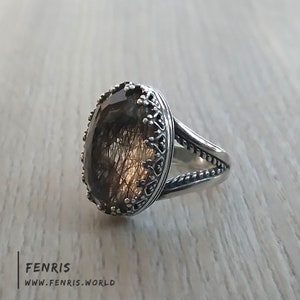 May include: A silver ring with a large oval-shaped gemstone. The gemstone is clear with brown and gold inclusions. The ring has a detailed ornate design around the gemstone.