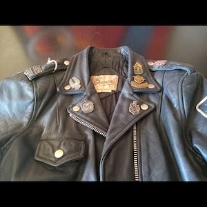 May include: Black leather motorcycle jacket with a zipper closure and multiple pins and patches. The jacket has a label that reads "Bennmans Fine Leather Outerwear".