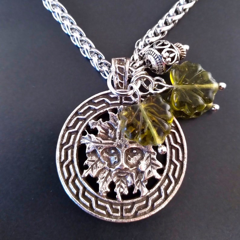 May include: A silver necklace with a circular pendant featuring a green man face surrounded by leaves. The pendant is attached to a silver chain with a small green glass leaf charm.