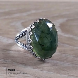 May include: A silver ring with a large, green, faceted gemstone. The gemstone has a unique, mossy appearance. The ring has a decorative, ornate band.