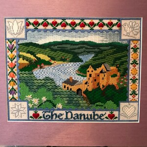 May include: A cross-stitch embroidery of a landscape with a castle and river. The embroidery is framed and has a border of flowers and hearts. The text "The Danube" is stitched below the image.
