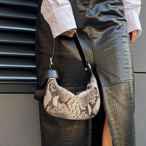 May include: A long black skirt with a crocodile-embossed texture and a front slit, paired with a white button-down shirt, a small crescent-shaped handbag featuring a grey and black snakeskin pattern, and black thong-style platform sandals.