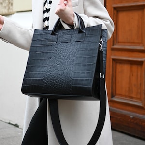 Può includere: Una borsa tote nera con finitura testurizzata effetto coccodrillo, dotata di due manici superiori e una tracolla nera rimovibile. La borsa ha una forma rettangolare e una piccola etichetta rettangolare del marchio centrata sul pannello frontale.