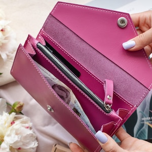 May include: A magenta leather wallet is open, revealing a zippered compartment and several pockets. The wallet is held open, showcasing its interior. White stitching accents the edges. A bill with Ukrainian text is visible.