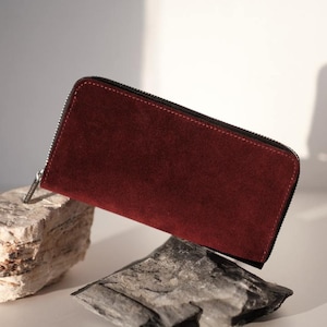 Puede incluir: Una cartera rectangular confeccionada en piel de ante color burdeos intenso, con un cierre de cremallera negro que recorre los bordes superior y lateral. La cartera tiene una textura mate y lisa con costuras precisas en todo el perímetro.