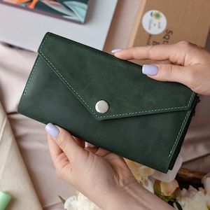 May include: A dark green leather wallet with a flap closure and a silver button. The wallet is held by hands with light purple nail polish. The stitching is visible around the edges. The wallet is on a soft, light pink surface.