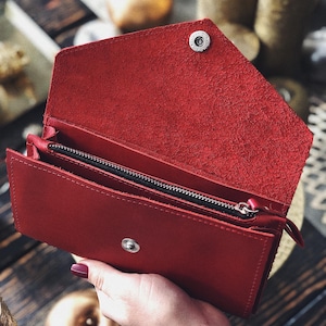 May include: A bright red envelope-style wallet featuring a textured interior flap, a central zippered compartment with a black zipper track, and a silver-toned metal snap button closure on the front exterior and the underside of the flap.