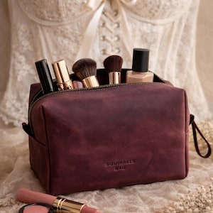 May include: A deep burgundy leather cosmetic bag with a zipper closure and the text SQUIRRELS BAG embossed on the front. The bag is filled with makeup brushes and bottles, while a lipstick, a small compact, and a square eyeshadow pan lie nearby.