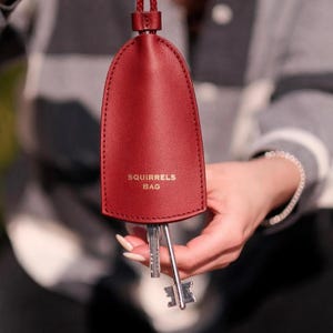 Könnte beinhalten: Ein weinroter Leder-Schlüsselhalter mit dem weißen Text "SQUIRRELS BAG". Der Schlüsselhalter ist oval und hat eine passende weinrote Kordel. Mehrere Schlüssel sind sichtbar, die an der Unterseite des Halters hängen.