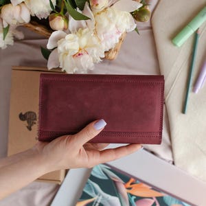 May include: A burgundy leather wallet with a stitched border is held. The rectangular wallet has a fold-over closure. The background includes a floral arrangement of white and pink peonies, a notebook, and stationery.