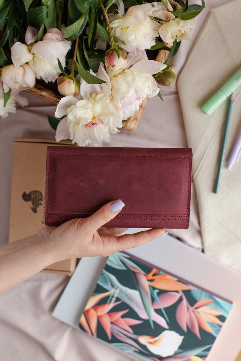 May include: A burgundy leather wallet with a stitched border is held. The rectangular wallet has a fold-over closure. The background includes a floral arrangement of white and pink peonies, a notebook, and stationery.