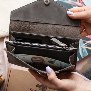 May include: A grey leather wallet with a snap closure, open to show a zipped compartment and banknotes. The wallet is held open, displaying the interior. The brand name "Squirrels Bag" is visible on a box in the background.