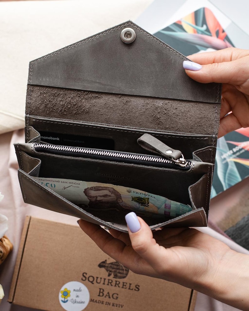 May include: A grey leather wallet with a snap closure, open to show a zipped compartment and banknotes. The wallet is held open, displaying the interior. The brand name "Squirrels Bag" is visible on a box in the background.