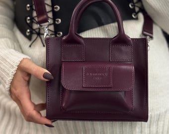 May include: A small, burgundy leather handbag with a top handle and a detachable shoulder strap. The bag features a front pocket with embossed text. The bag is held by a person.