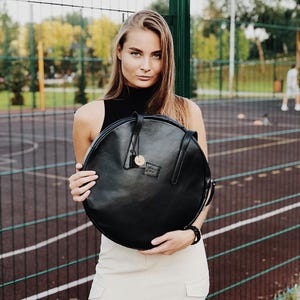 May include: A circular black leather bag with a smooth finish, featuring two thin shoulder straps, a small round hanging tag, and a rectangular brand patch on the front. The bag has a zipper closure along the top edge and a structured, flat design.