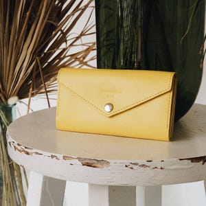 May include: A yellow leather wallet with a flap closure and a silver snap. The wallet has the words "Squirrels Bag" embossed on the front. It is sitting on a white, round stool. The background includes dried plants and a green vase.
