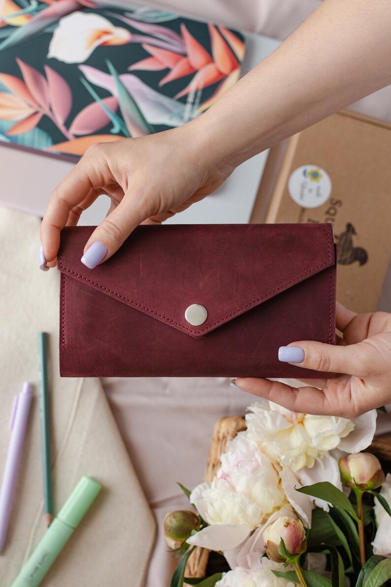 May include: A burgundy leather wallet with a flap closure and a silver button. The wallet is held by a person. The background includes a floral patterned laptop, pens, and flowers.