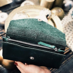 May include: Open, dark green leather wallet with a silver zipper and snap closure. The wallet is open, revealing the interior. The leather has a textured appearance. The wallet is held by a person.