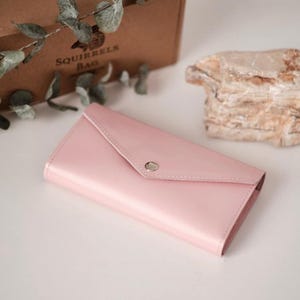 May include: A pale pink envelope-style wallet with a silver snap closure. The wallet is made of a glossy material and is lying on a white surface. A brown box with the text "SQUIRRELS BAG" is in the background.