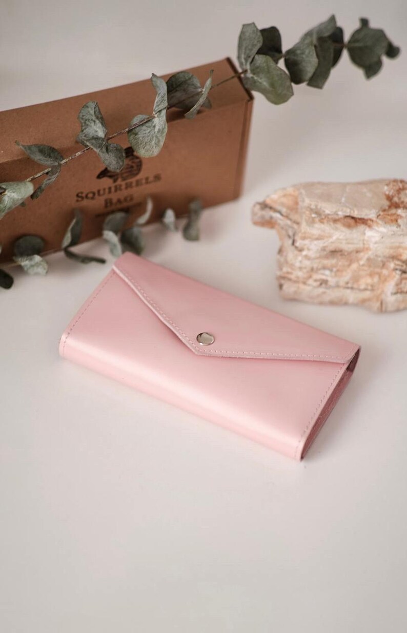 May include: A pale pink envelope-style wallet with a silver snap closure. The wallet is made of a glossy material and is lying on a white surface. A brown box with the text "SQUIRRELS BAG" is in the background.
