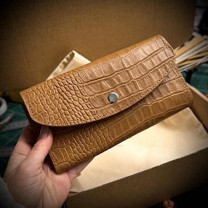 May include: A tan wallet with a textured crocodile skin pattern, featuring a curved flap closure secured by a single silver-toned metal snap button. The wallet has visible stitching along the edges of the flap and a rectangular body shape.