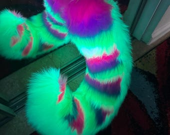 May include: A long, curved faux fur tail featuring a vibrant pattern of neon green, bright pink, and deep purple stripes. The plush, shaggy texture of the synthetic fur creates a dense, voluminous appearance with a distinct, multi-colored gradient.
