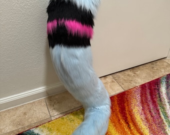 May include: A fluffy, light blue faux fur tail with a curved shape. The tail features a black and hot pink stripe near the top. The tail is designed to be worn as a costume accessory.