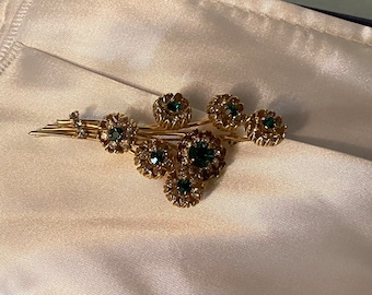 May include: A gold-toned brooch with a cluster of green and clear rhinestones arranged in a floral design.