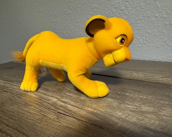 May include: A plush toy lion cub in a walking pose. The lion is primarily yellow with a tan belly and a fluffy tail. The ears are black-lined, and the eyes are detailed with brown and yellow. The toy is on a wooden surface.