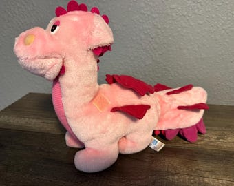 May include: A plush dragon toy in a light pink color with a textured pink chest panel. It features dark pink spikes along its back and tail, dark pink wings, and a yellow patch on its snout. The dragon has a soft, fuzzy texture throughout.