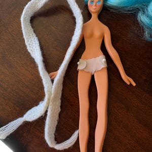 May include: A vintage doll with blue hair and a white knitted scarf. The doll is wearing a light pink bottom with pearl accents and pink shoes. The doll's skin tone is peach.