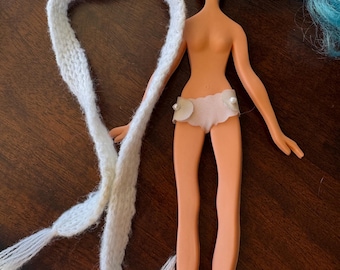May include: A vintage doll with blue hair and a white knitted scarf. The doll is wearing a light pink bottom with pearl accents and pink shoes. The doll's skin tone is peach.