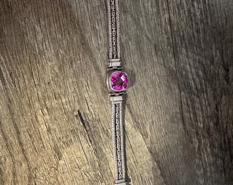 May include: A silver bracelet featuring a woven chain design with a central square-cut, faceted pink gemstone. The bracelet includes a metallic clasp mechanism at one end and a decorative metal end piece at the other, showcasing intricate detailing.