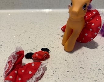 May include: A vintage toy pony with a yellow body, purple hair, and a red polka dot dress. The pony wears a matching headband with black ears. A separate dress and headband are also visible.