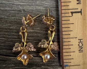 May include: A pair of gold-tone drop earrings featuring a floral design with small leaves and a single white pearl at the base of each. A wooden ruler is placed next to the earrings, showing they measure approximately 0.75 inches in length.