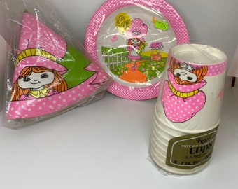 May include: A set of pink and green party supplies featuring a girl with a basket of flowers. The set includes paper plates, cups, and party hats. The plates have a pink border with white polka dots and the text "Happy Birthday". The cups have a pink and white polka dot pattern. The party hats are pink with white polka dots and a green ribbon.