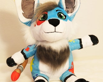 May include: A blue and white plush toy with paint splatters and a paintbrush tail. The toy has grey ears, a grey beard, and black paws with blue paw pads.
