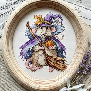 Könnte beinhalten: Ein ovaler Stickrahmen aus Holz mit einer detaillierten Stickerei einer Maus, die als Hexe verkleidet ist. Die Maus trägt einen violetten Hut mit einem Ahornblatt, einen violetten Umhang und hält einen kleinen orangefarbenen Kürbis sowie einen Besen.