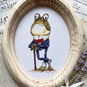 May include: A cross-stitch embroidery of a frog wearing a blue suit and a red bow tie, leaning on a cane. The embroidery is framed in an oval light-colored wooden frame, set against a background of sheet music and a sprig of dried lavender.