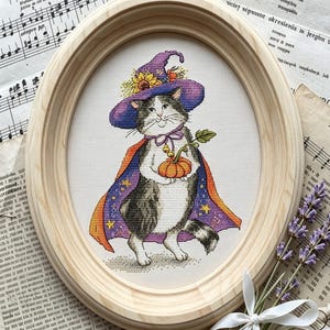 Könnte beinhalten: Eine Kreuzstichstickerei in einem ovalen Holzrahmen, die eine grau-weiße Katze mit einem violetten Hexenhut, verziert mit Sonnenblumen, und einem violetten Umhang mit gelben Sternen zeigt, die einen kleinen orangefarbenen Kürbis mit grünem Stiel hält.