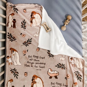 Op de afbeelding: Een zachte, beige fleece deken met een bloemenpatroon en illustraties van Maria en Jezus. De deken heeft de tekst "Maar Maria bewaarde al deze dingen in haar hart en overdacht ze. Lk 2:19".
