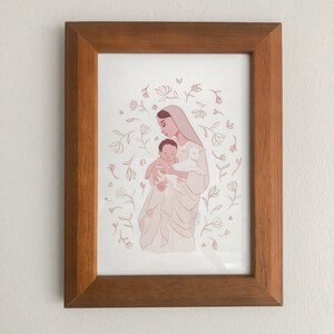 May include: A framed illustration of a woman holding a baby and a lamb. The woman is wearing a white robe and has a halo. The baby is wrapped in a white blanket. The lamb is white and has a pink nose. The background is a light pink with white flowers.