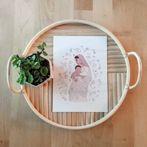 May include: A white print with a pink illustration of a woman holding a baby. The woman is wearing a veil and the baby is wearing a white garment. The illustration is surrounded by a circle of pink flowers and leaves.