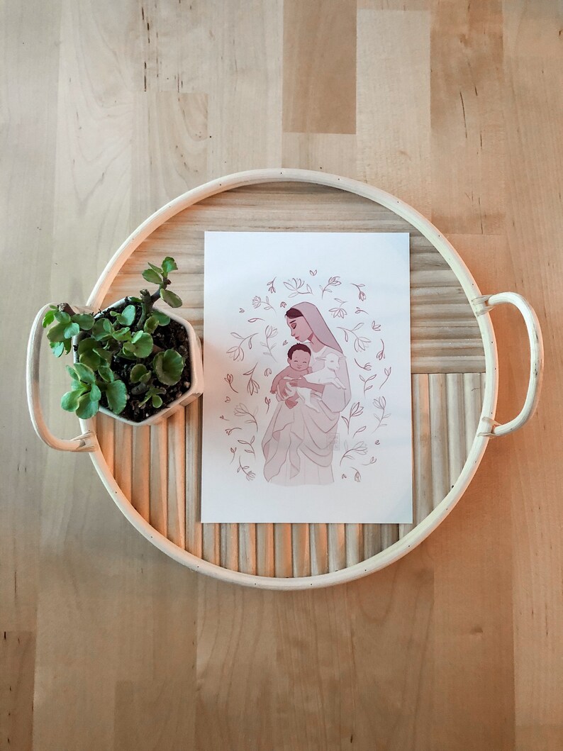 May include: A white print with a pink illustration of a woman holding a baby. The woman is wearing a veil and the baby is wearing a white garment. The illustration is surrounded by a circle of pink flowers and leaves.