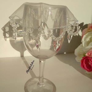 Può includere: Un bicchiere di vino in vetro trasparente con una copertura in tulle bianco e perline di cristallo. La copertura ha un bordo argentato.