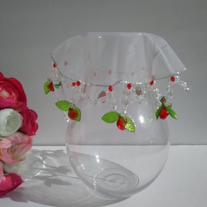 Può includere: Un vaso di vetro trasparente con un bordo di tulle bianco e perline rosse e verdi. Il vaso è decorato con piccole perline rosse e verdi che assomigliano a ciliegie.