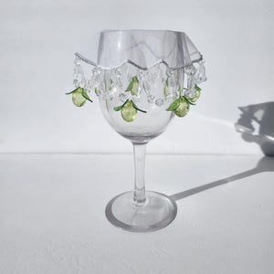 May include: A clear wine glass with a decorative white fabric cover. The cover is trimmed with clear beads and green fruit-shaped charms. The glass sits on a white surface, casting a shadow.