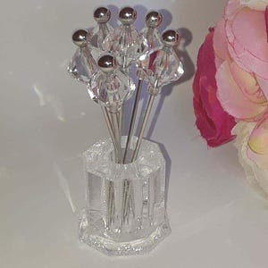 May include: A set of six decorative cocktail picks with clear, faceted crystal-like tops and silver stems, displayed in a clear, hexagonal glass holder. The background is a soft white with a hint of pink floral accents.
